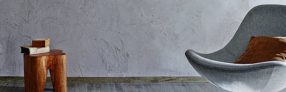 Holzhocker und grauer Sessel vor grauer Lehm-Edelputz Wand